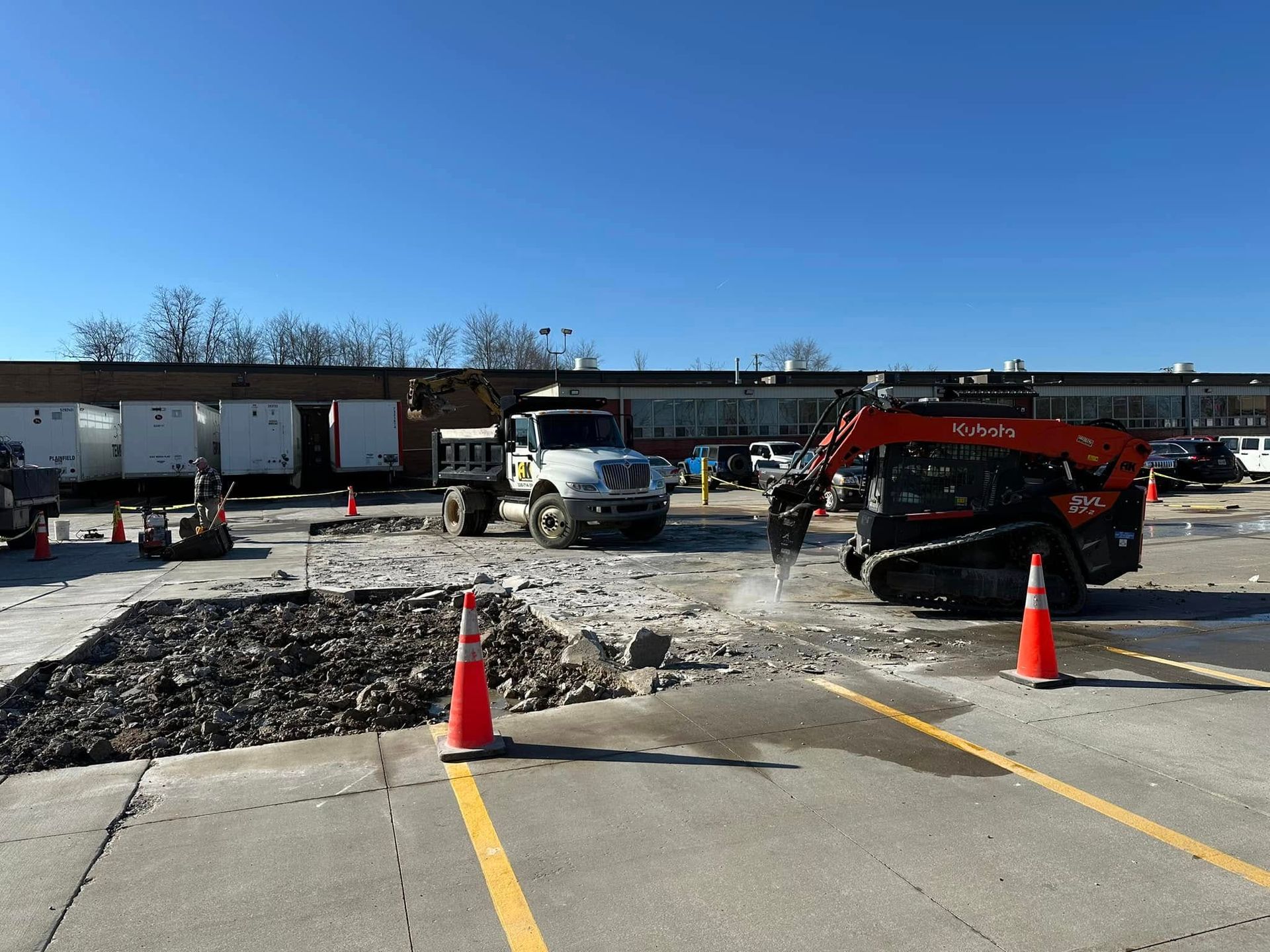 Construction site: a truck and excavator breaking pavement. Orange cones mark the work area; clear sky.