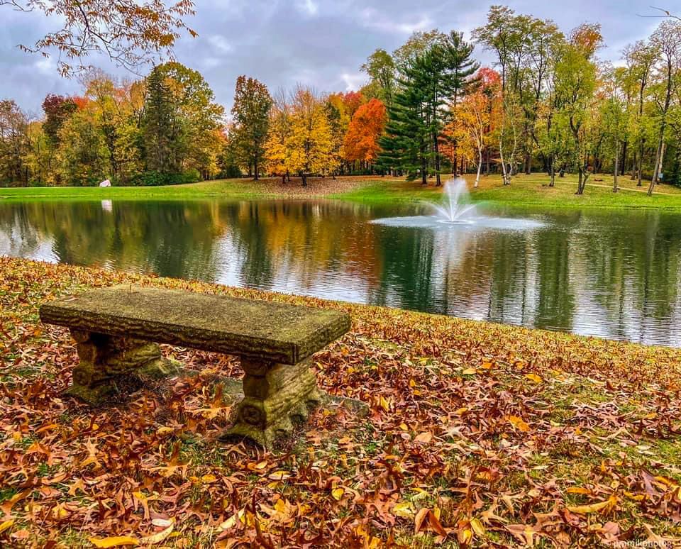 Stone bench by a pond with fountain, surrounded by colorful autumn trees and fallen leaves.