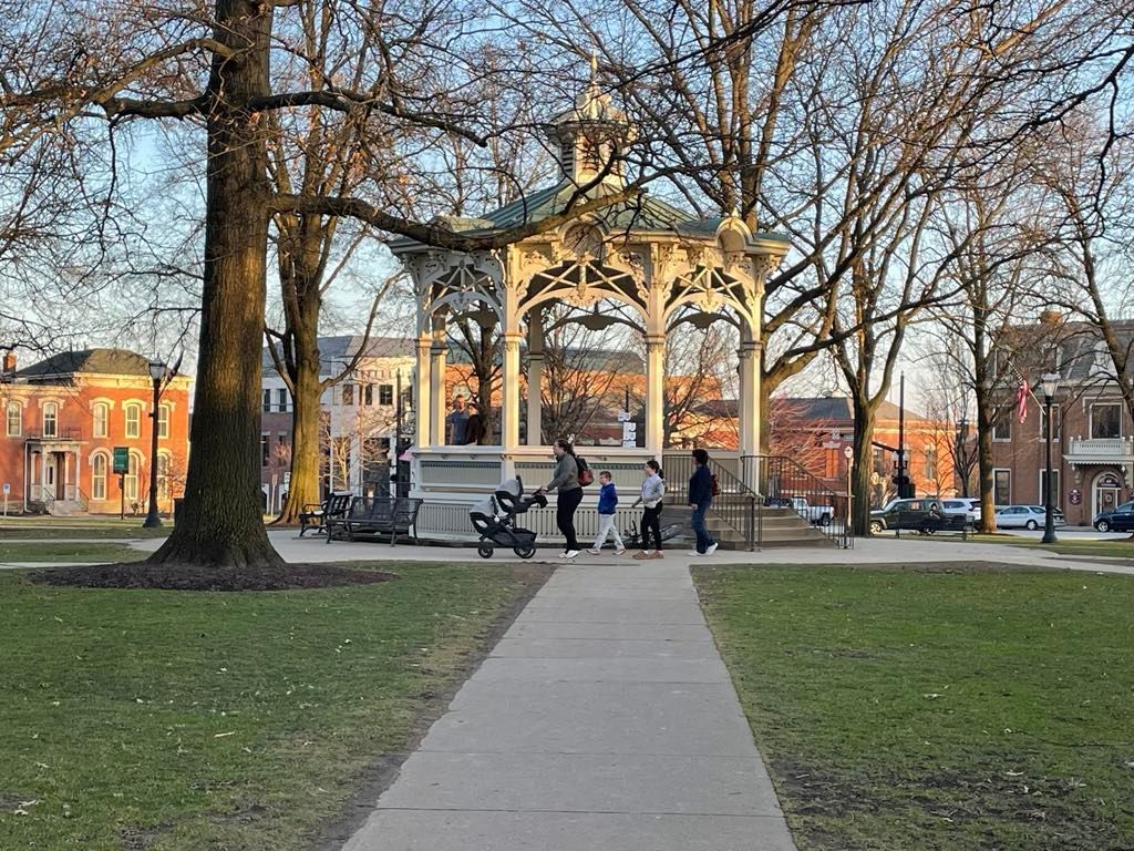 Gazebo in a park with several people standing nearby. Buildings and trees surround.