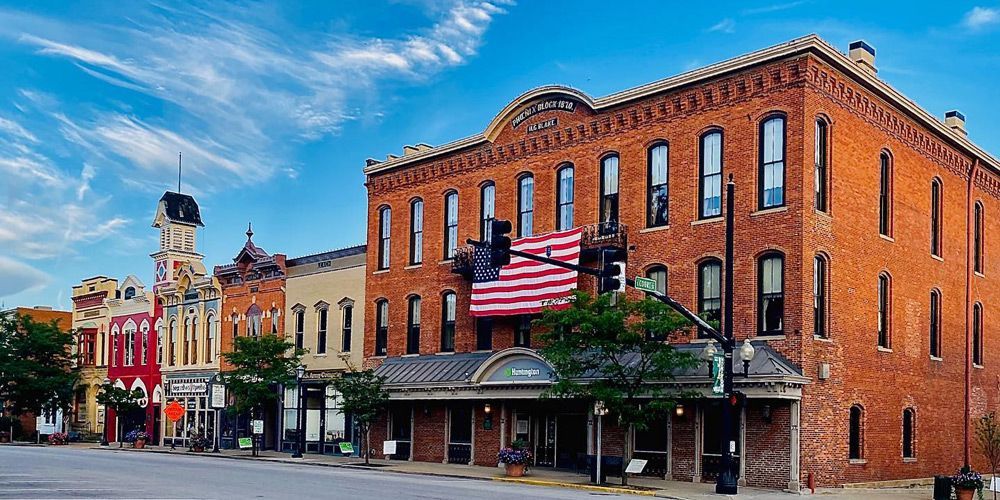 Buildings line a town street; brick buildings, one with an American flag. Blue sky.