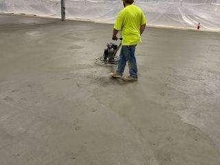 Worker using a power trowel on a concrete floor inside a building, wearing a yellow shirt and jeans.