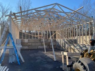 Wooden frame of a building under construction, with a partially built roof, concrete blocks, and a clear blue sky.