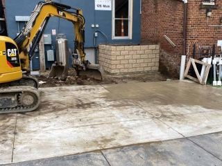 Yellow excavator at a construction site, next to a brick wall and a blue building.