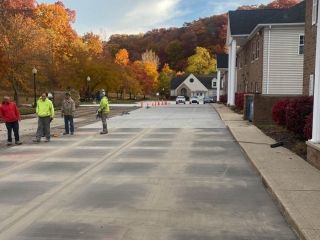 Workers on a newly paved road; trees in fall colors in the background; buildings on right side.