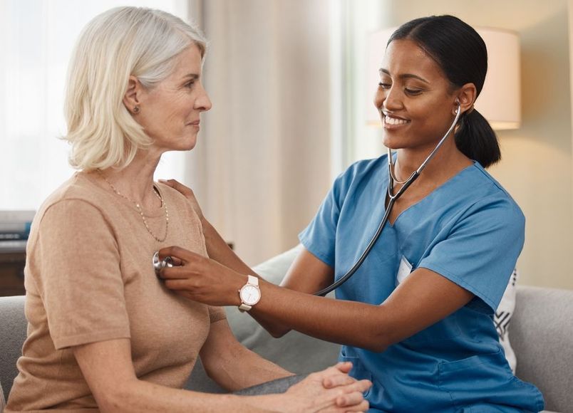 A healthcare worker in blue scrubs uses a stethoscope to examine a patient sitting on a couch.