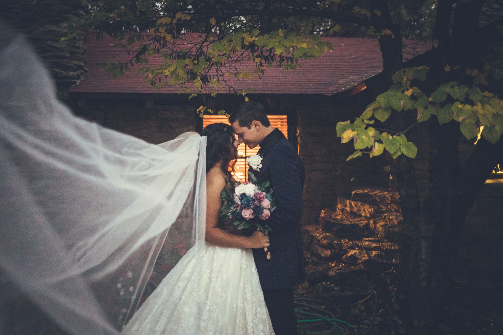 A bride and groom are kissing in front of a building with a veil blowing in the wind.