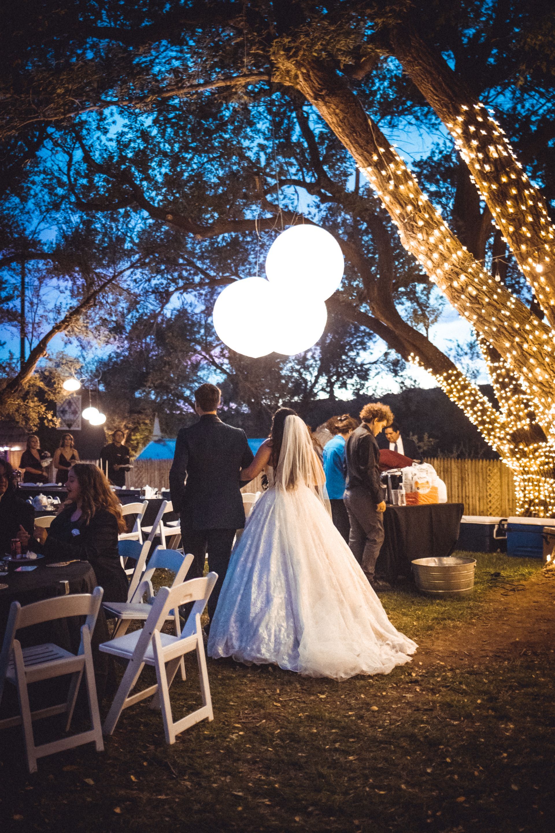 A bride and groom are walking under a tree with lights.