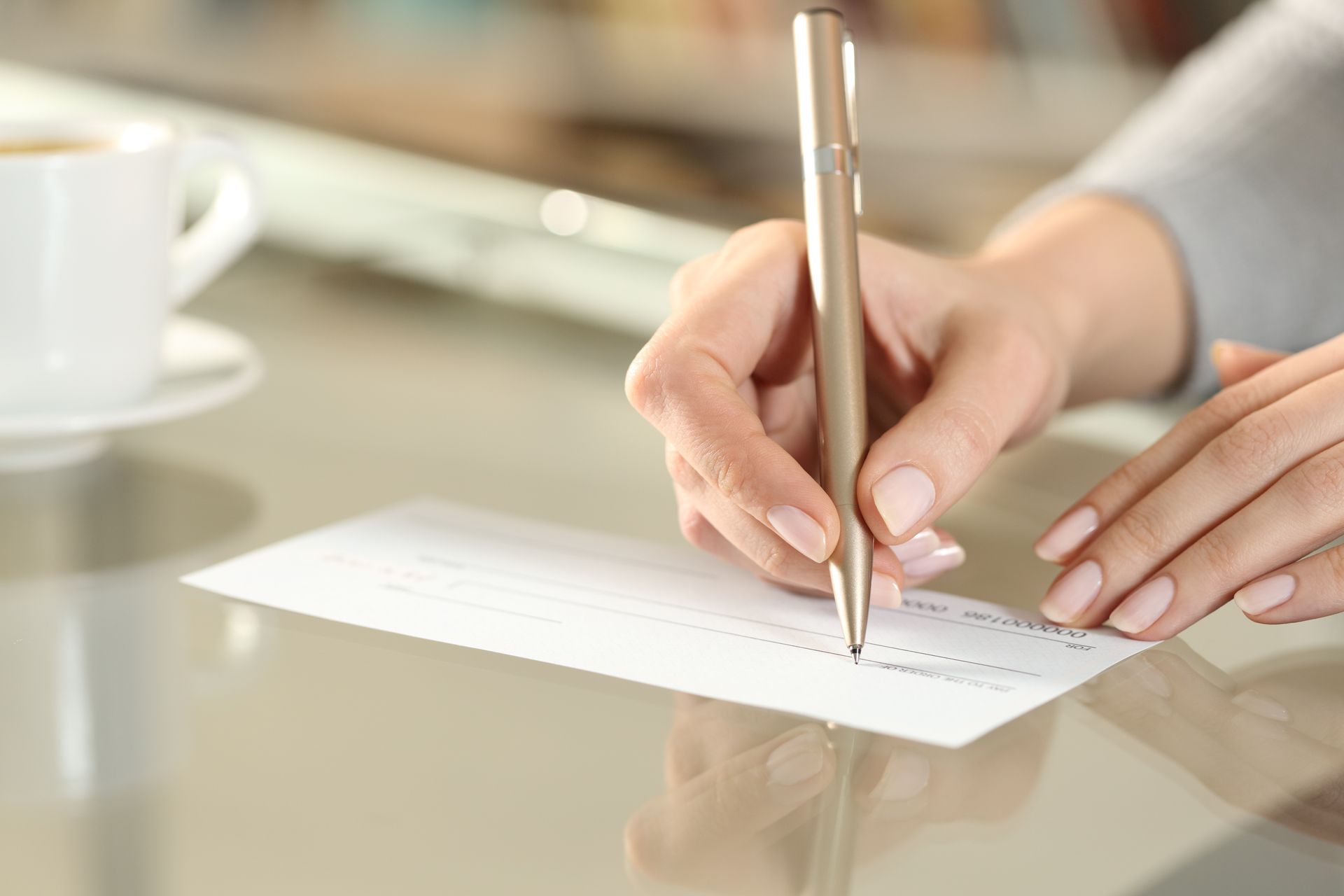 A person's hands writing with a pen on a blank check at a table, coffee cup in the background.