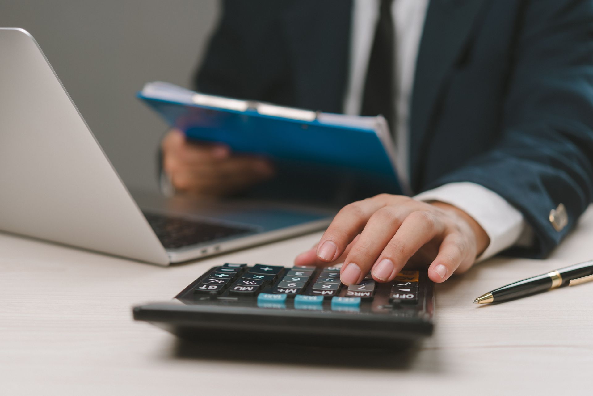 Person in suit using a calculator, laptop, and document on a desk.