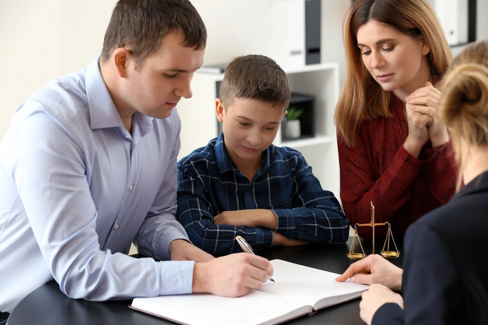 Family with lawyer at table, man signing document; scales of justice.