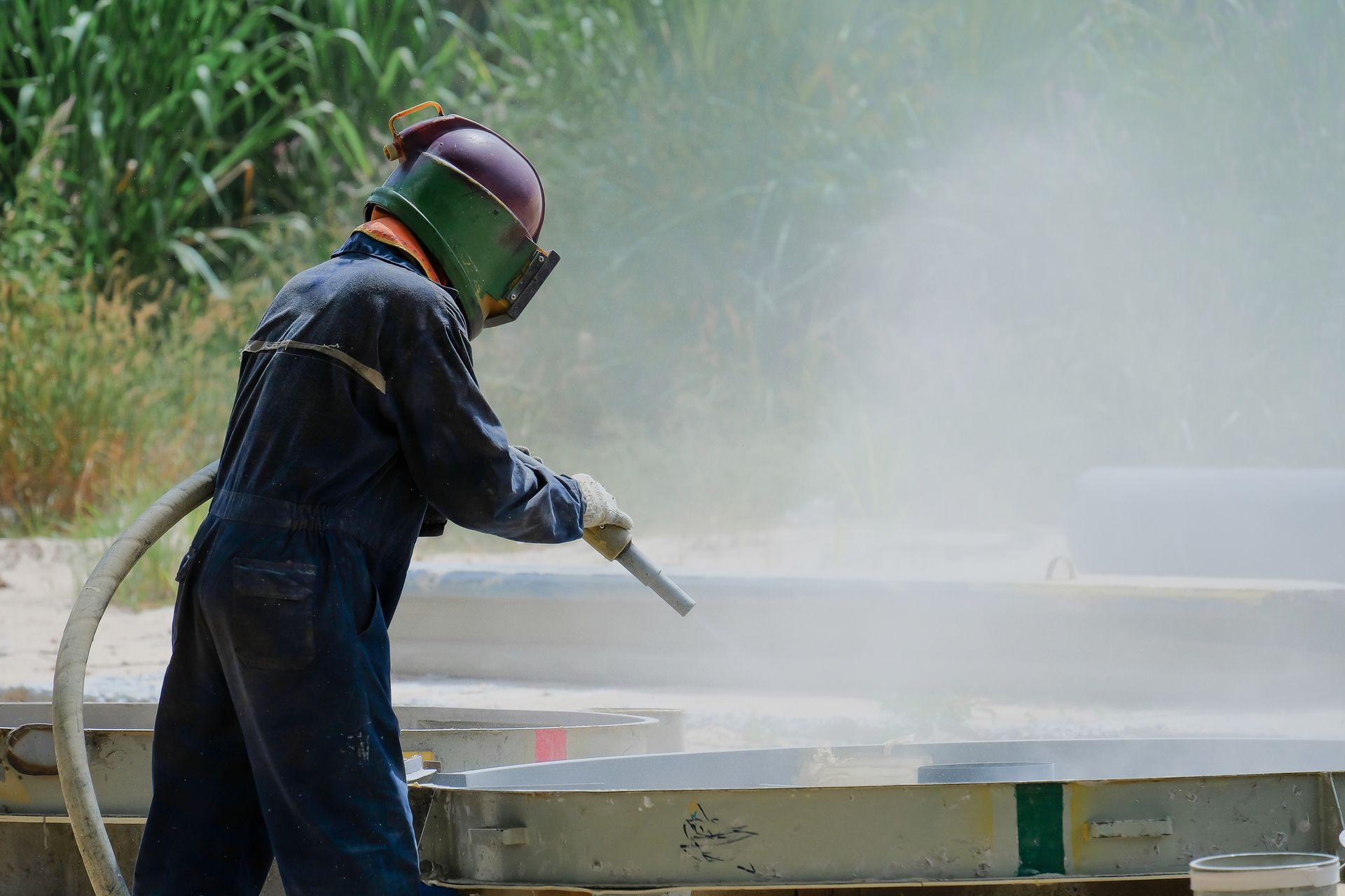 Person in protective gear blasting metal outdoors, creating a dust cloud.