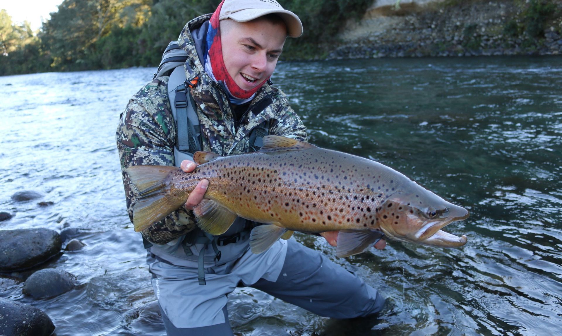 A man is kneeling in the water holding a large brown trout.