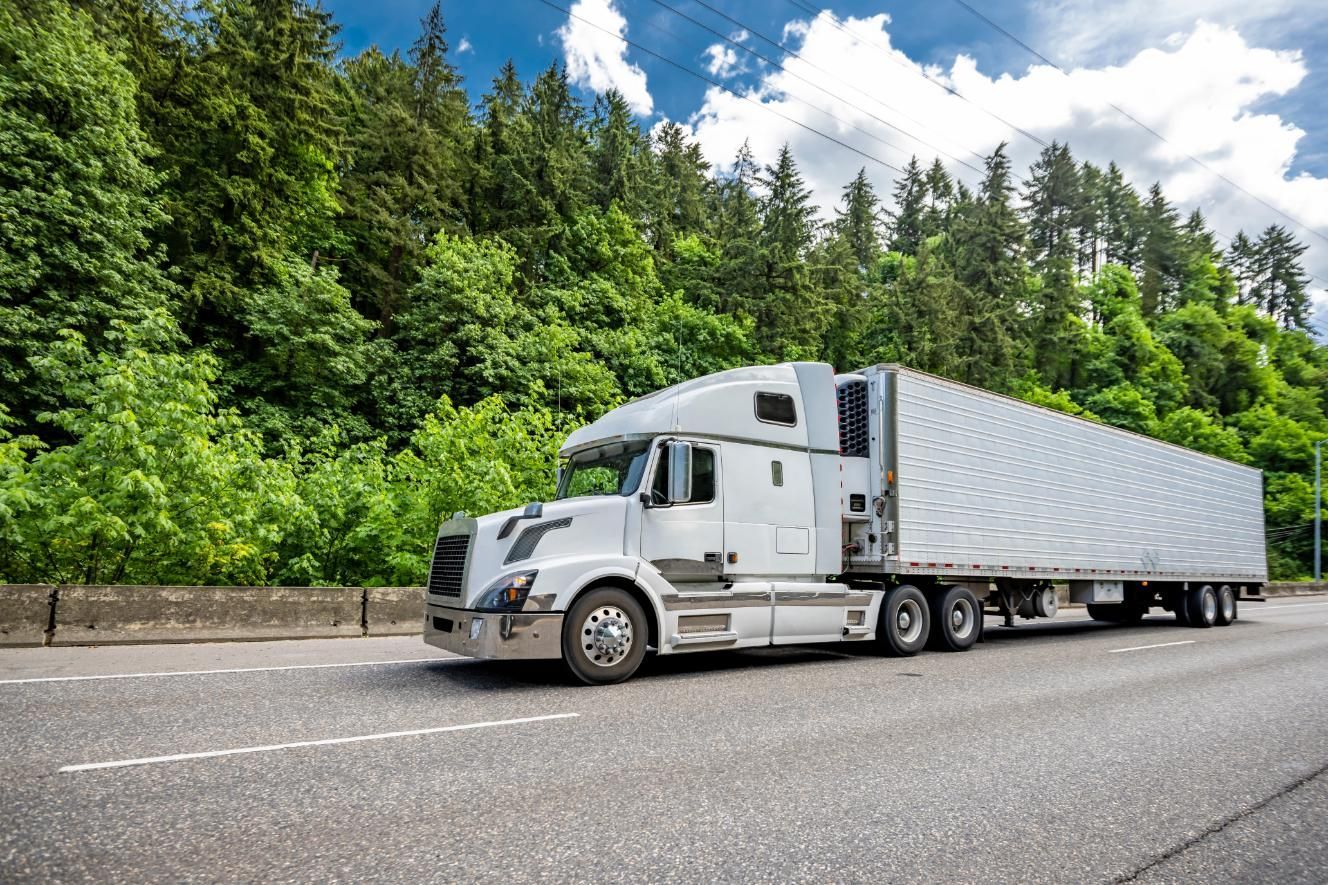 A white semi truck is driving down a highway next to a forest.