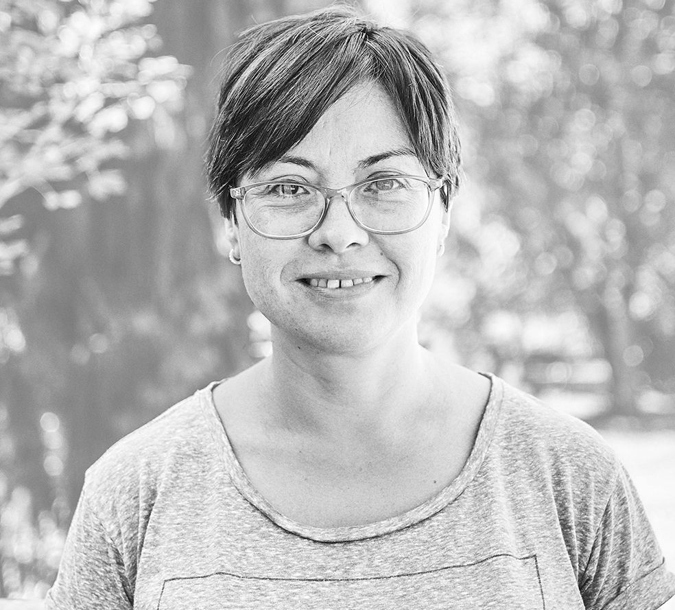 Eine Frau mit Brille und grauem Hemd lächelt auf einem Schwarzweißfoto.