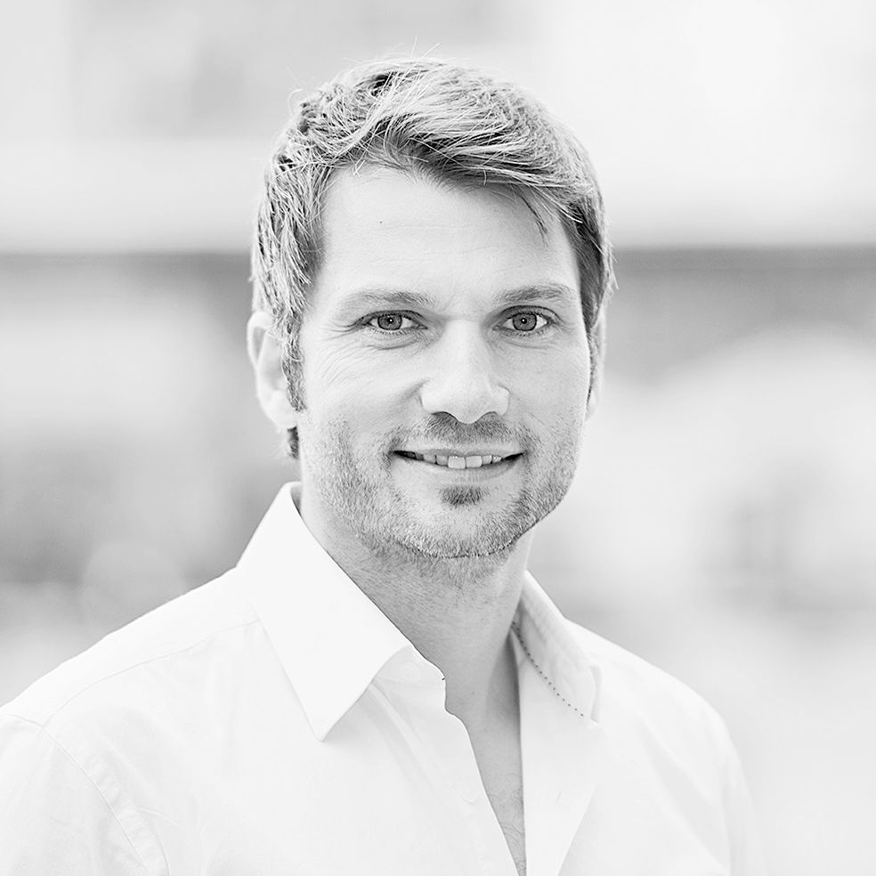 Auf einem Schwarzweißfoto trägt ein Mann mit Bart ein weißes Hemd und lächelt.