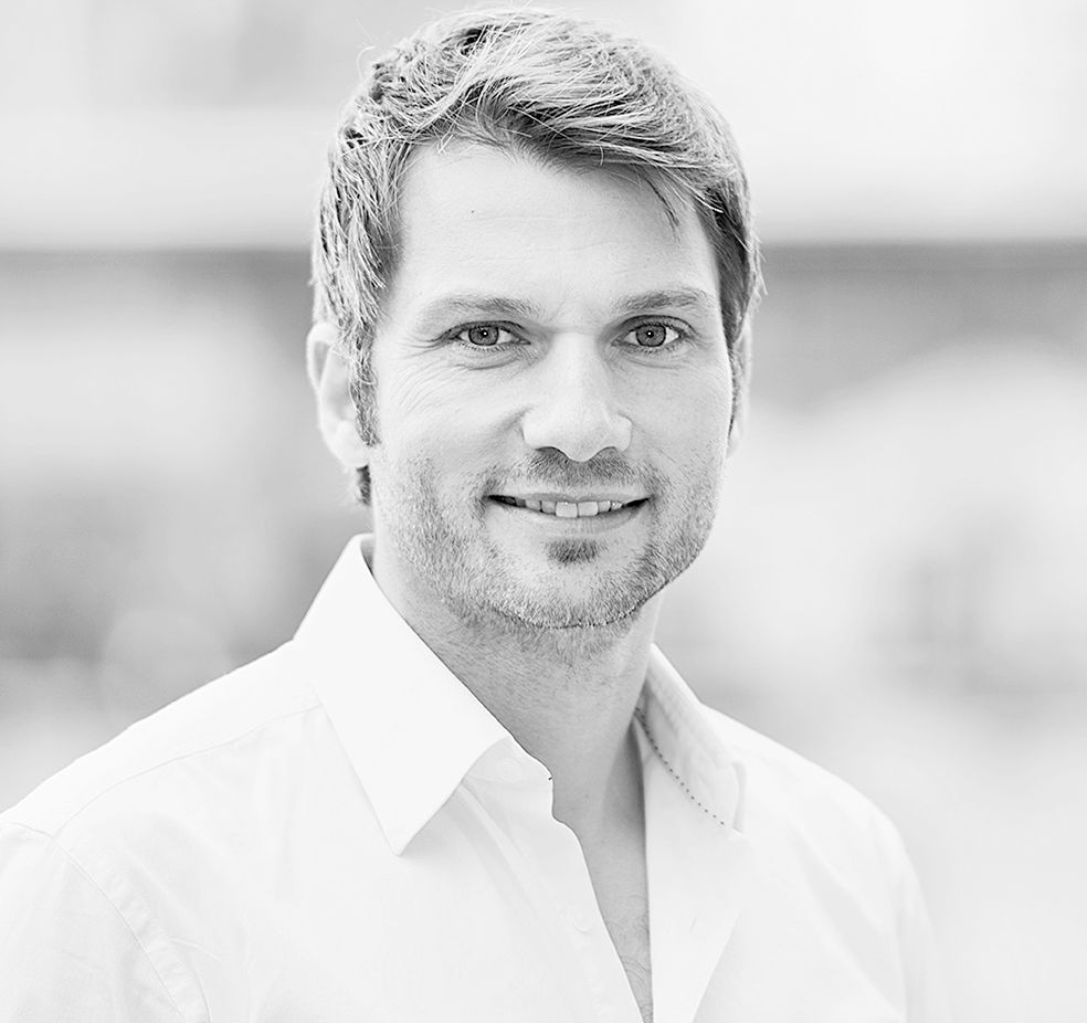 Auf einem Schwarzweißfoto trägt ein Mann mit Bart ein weißes Hemd und lächelt.