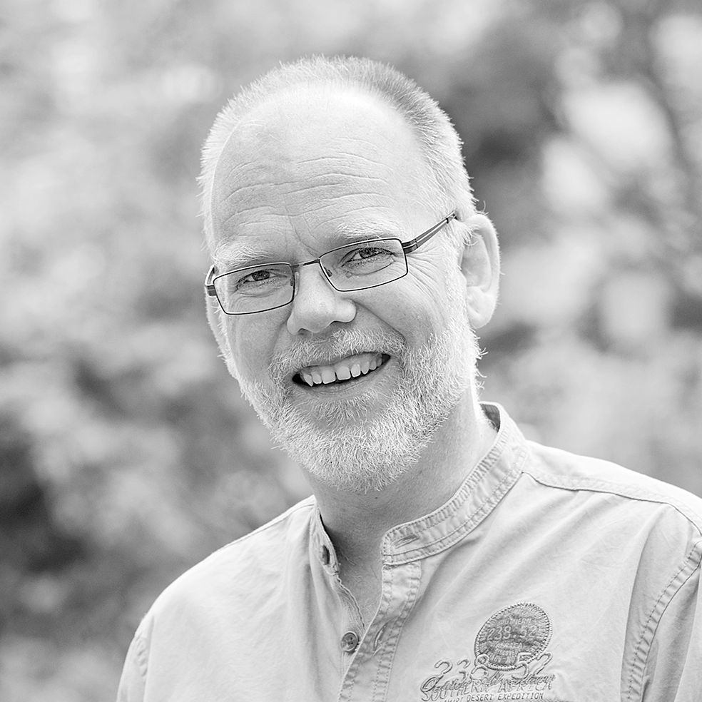 Ein Mann mit Bart und Brille lächelt auf einem Schwarzweißfoto.