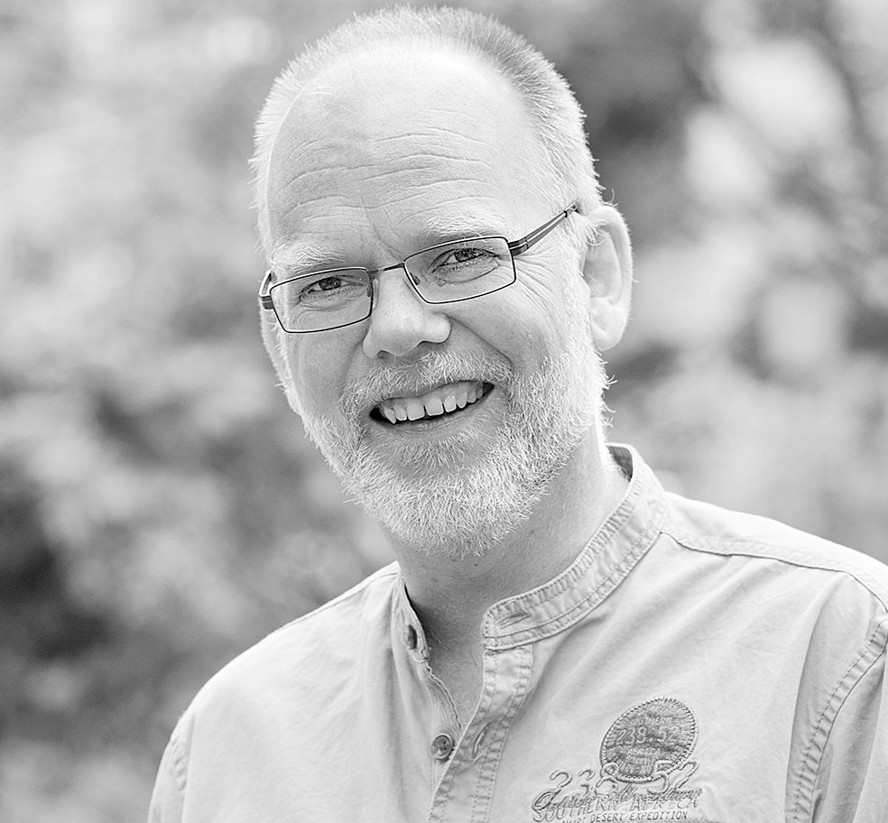 Ein Mann mit Bart und Brille lächelt auf einem Schwarzweißfoto.