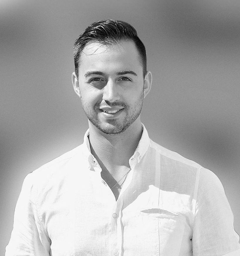 Auf einem Schwarzweißfoto trägt ein Mann mit Bart ein weißes Hemd und lächelt.