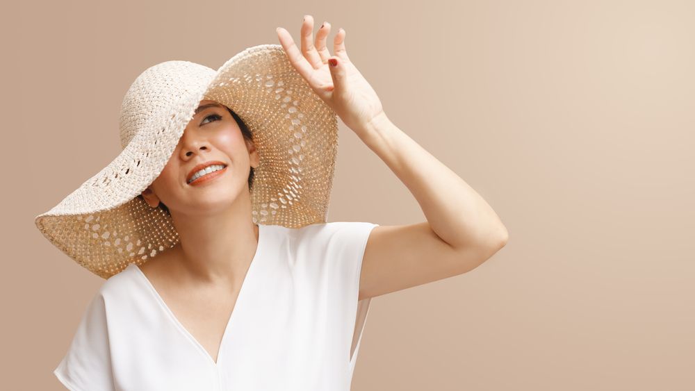 Woman wearing a wide-brimmed straw hat shields her eyes with her hand, smiling, against a neutral background.