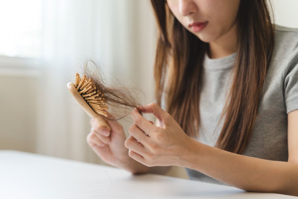 Woman examining hair loss on a wooden hairbrush. She's holding the brush, looking concerned, indoors, wearing a gray shirt.
