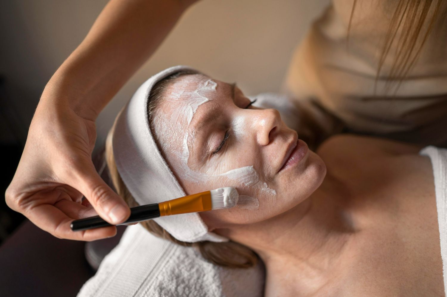 Woman receiving facial treatment in a spa setting; a cosmetologist uses a brush to apply a white mask to her face.