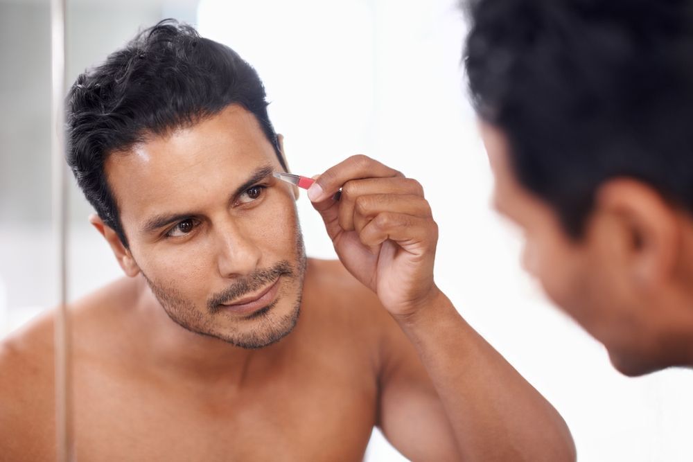 A man plucking his eyebrow in a mirror. He has dark hair and skin, and is shirtless.