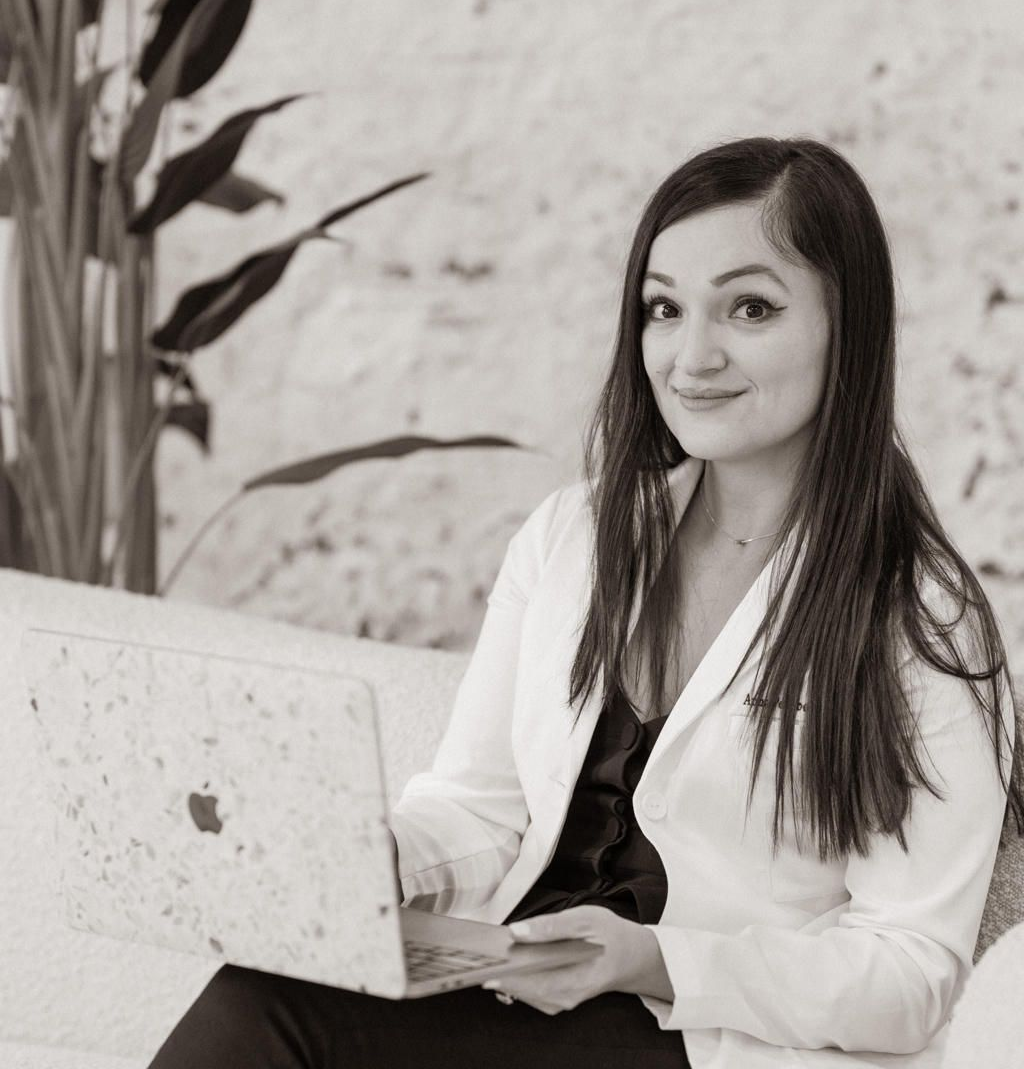 A person with long dark hair, wearing a white blazer, sits holding a textured laptop in a bright, modern office space.