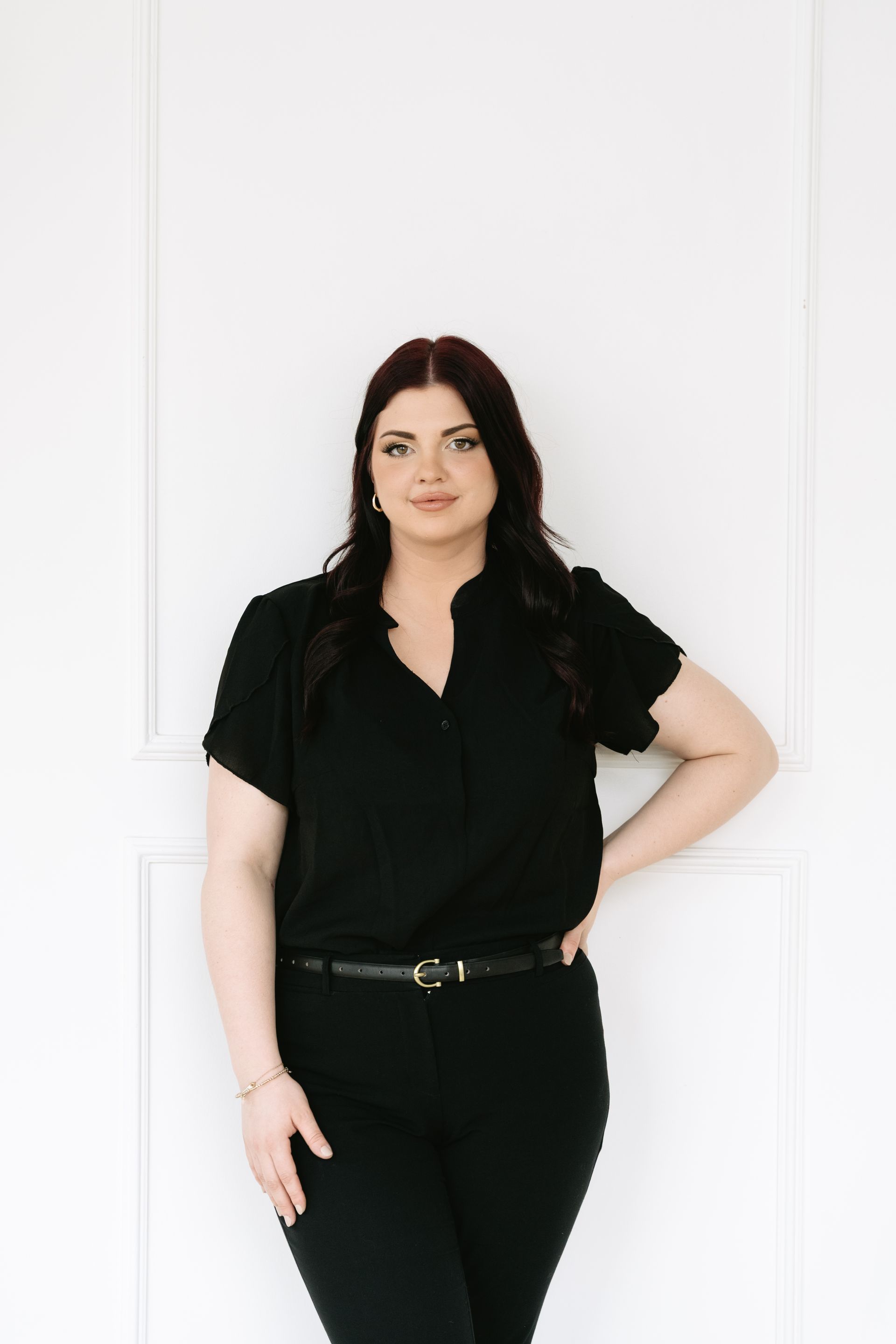 A woman in a black shirt and black pants is standing in front of a white wall.