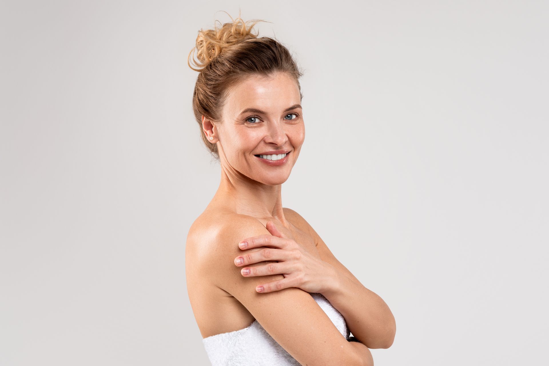 A woman wrapped in a white towel is smiling and touching her shoulder.