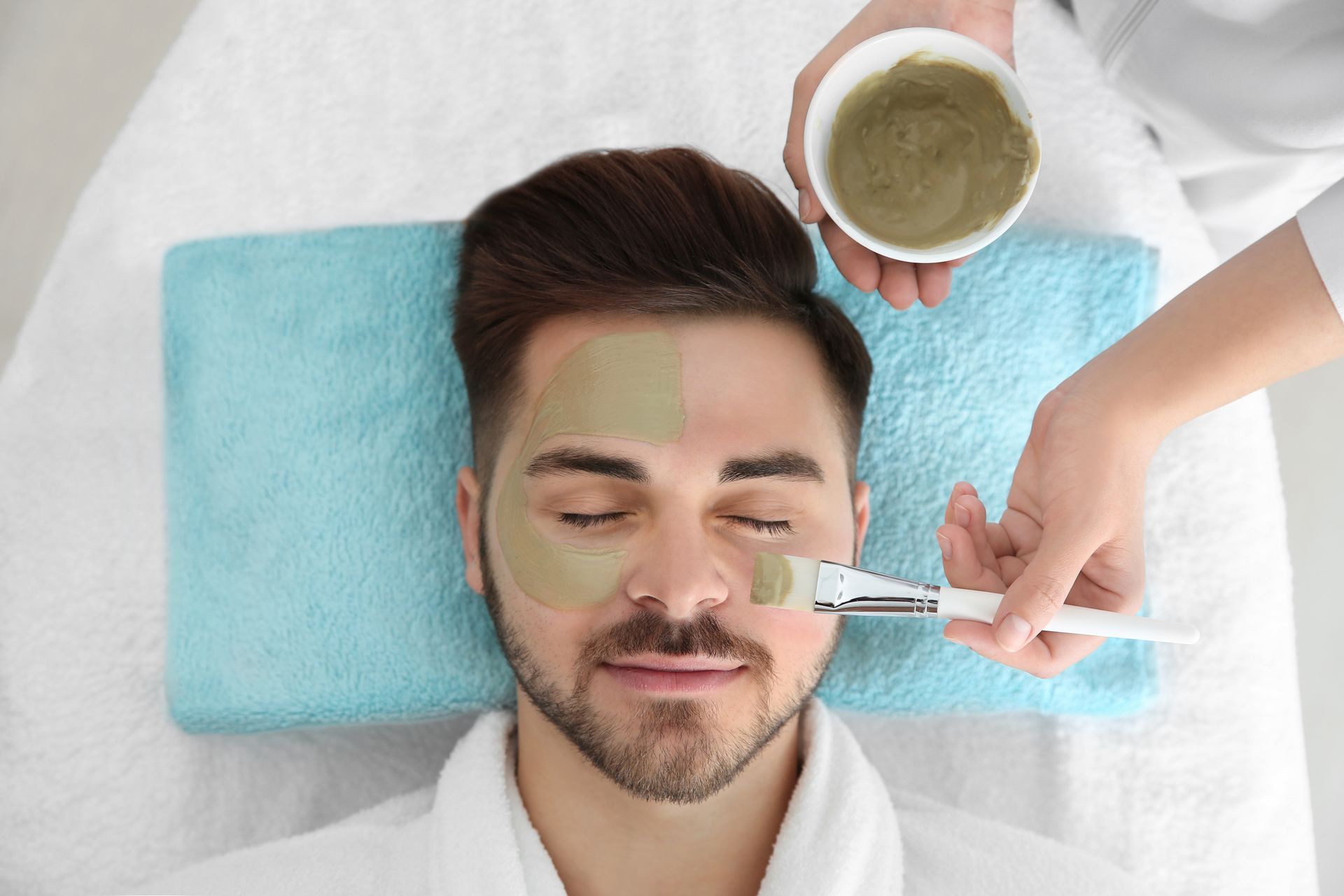 Man receiving a facial mask in a spa setting; someone applying mask with a brush.