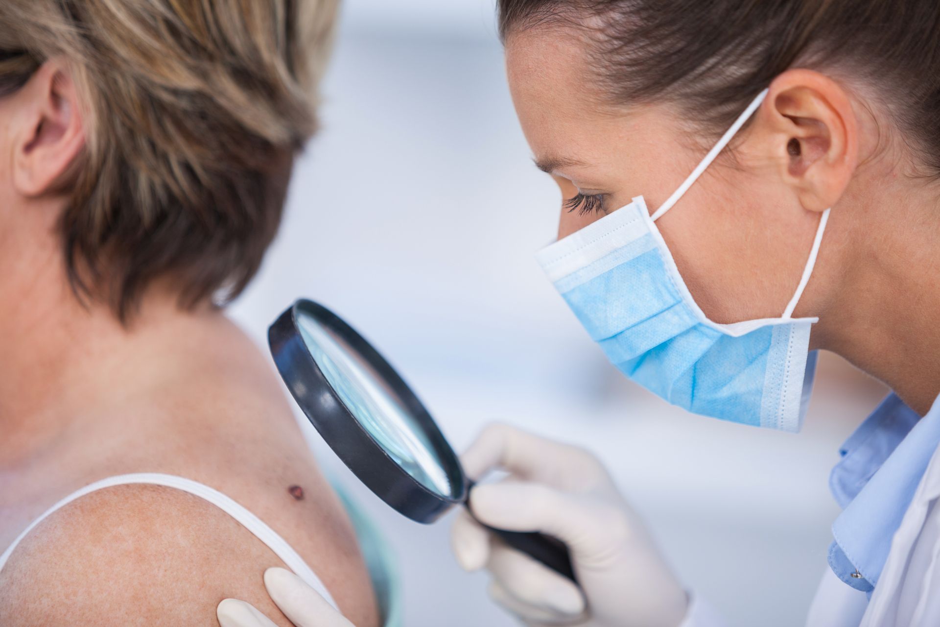 Dermatologist in mask examines a mole on a patient's shoulder with a magnifying glass.
