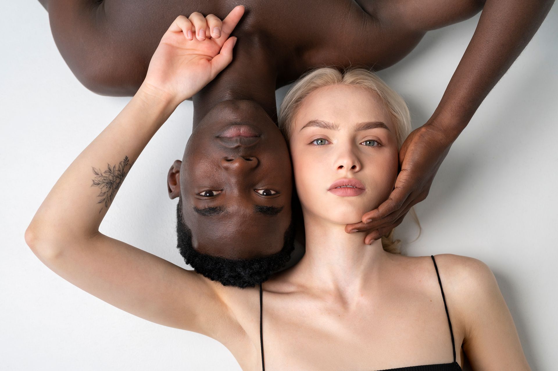 Two people, one with dark skin, the other with light skin, lying head-to-head. Hands touch their faces.
