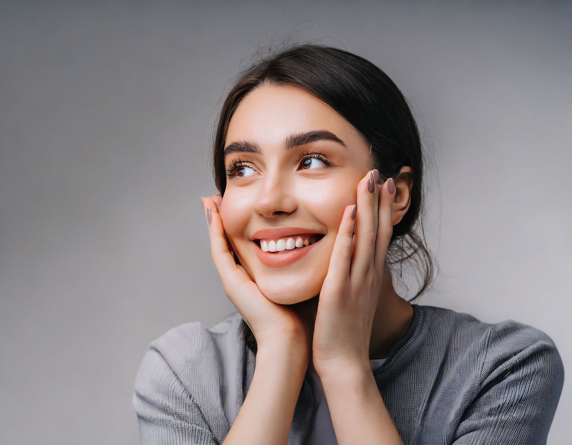 Smiling woman with hands on cheeks, looking upward, against a gray background.