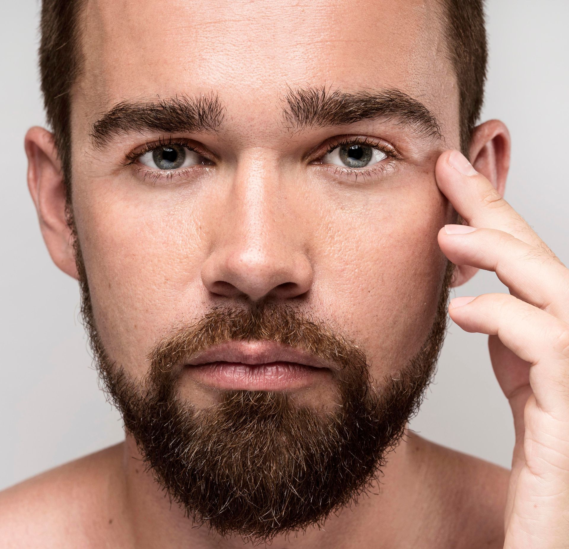 A man with a beard is touching his eye with his hand.