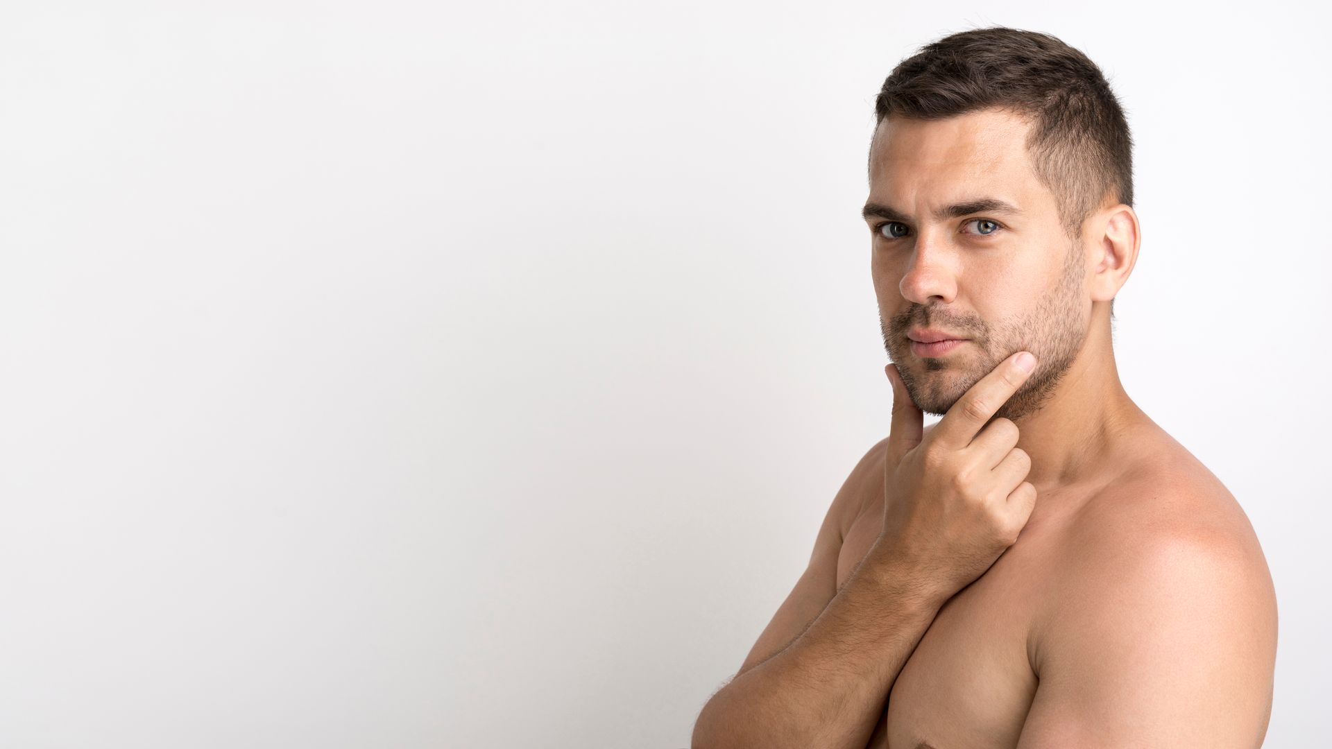 A shirtless man is holding his hand to his chin and looking at the camera.