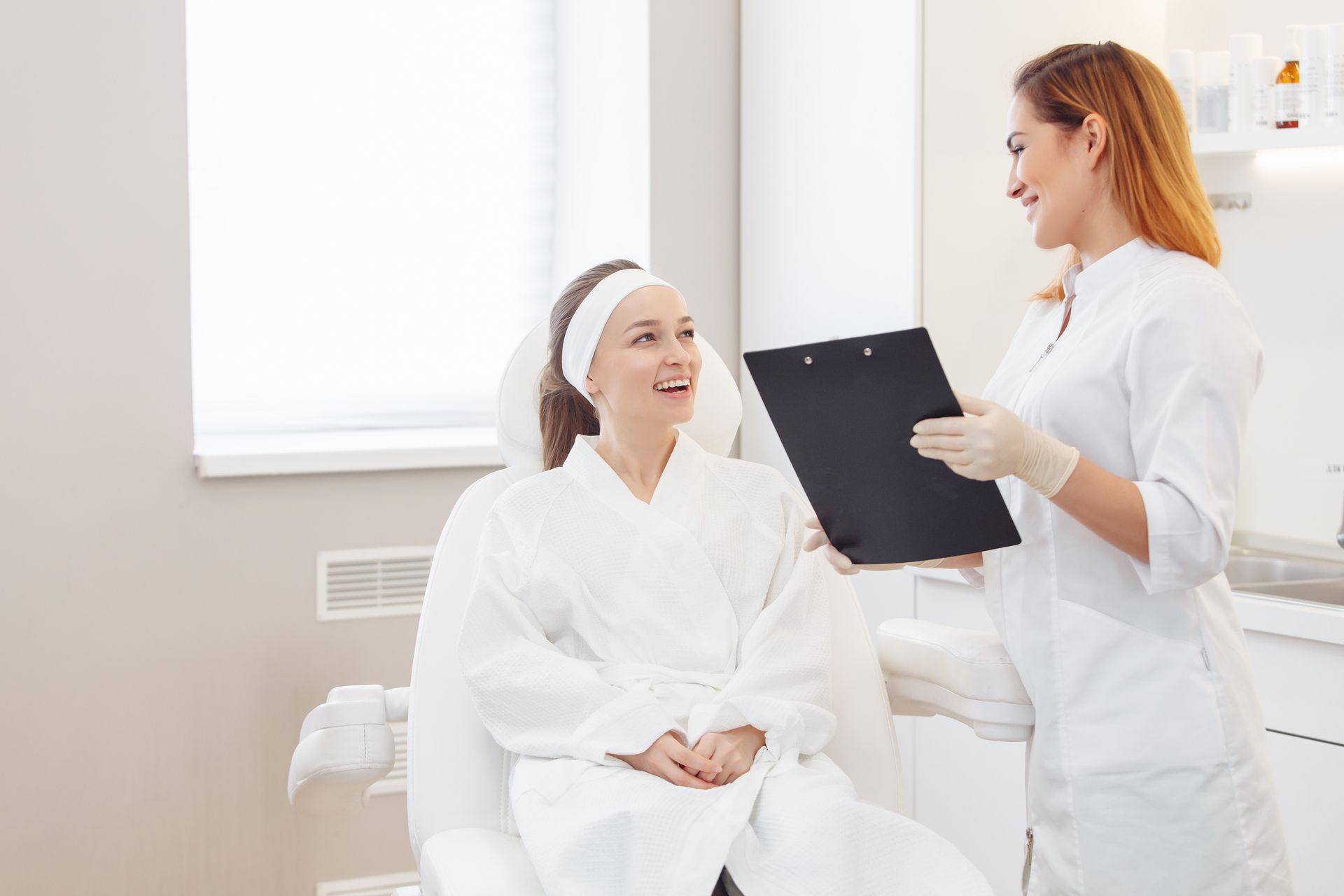 Woman in spa robe smiles at a professional holding a clipboard in a bright room.