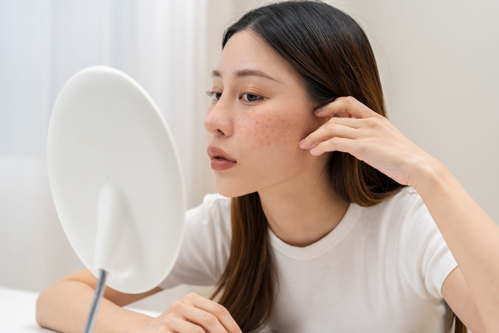 Woman looking in a mirror, examining skin. She has freckles on her cheeks and wears a white shirt.