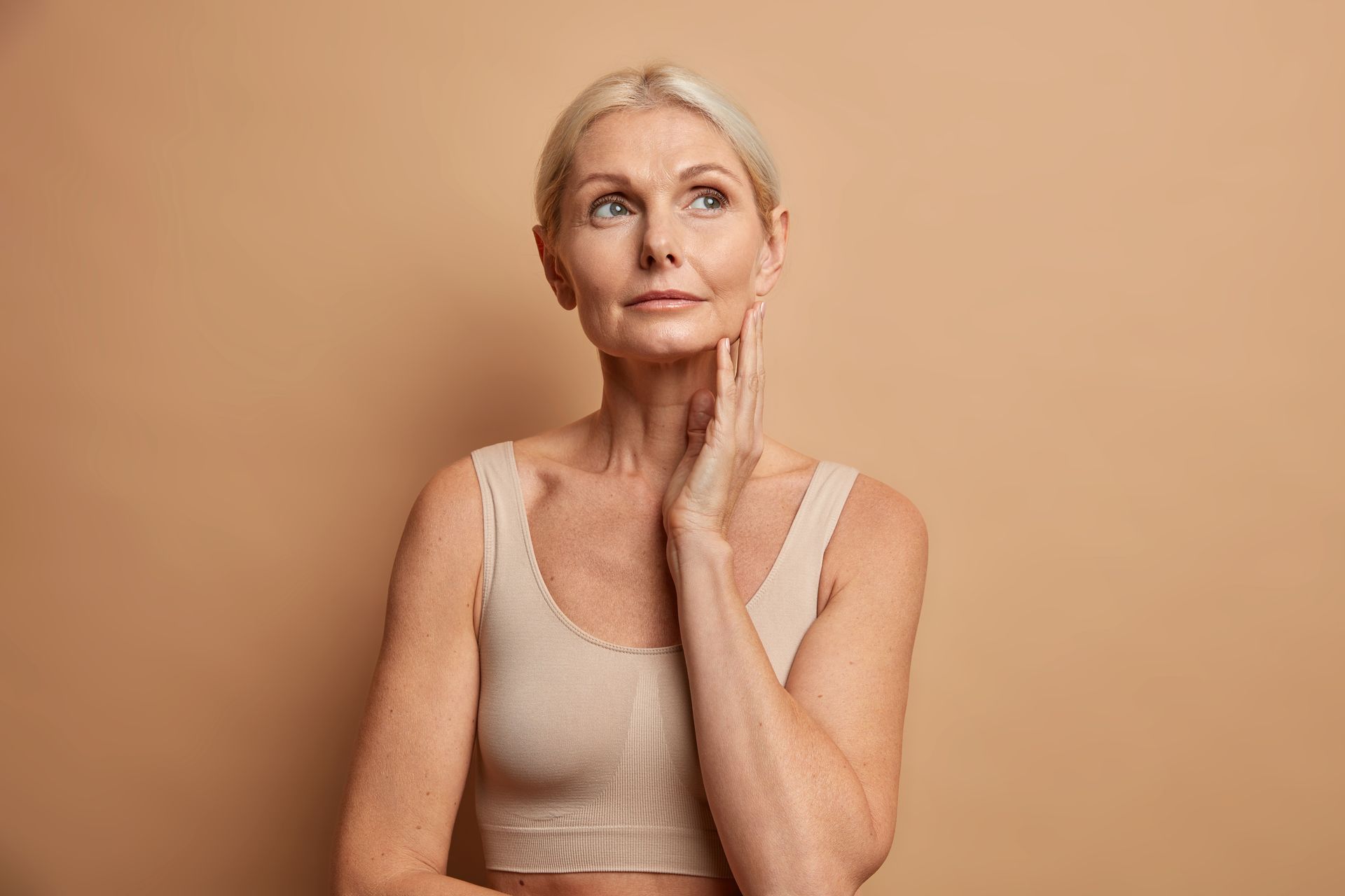 Woman with fair skin and short blonde hair, gazing upwards while touching her cheek, wearing a neutral-colored sports bra.