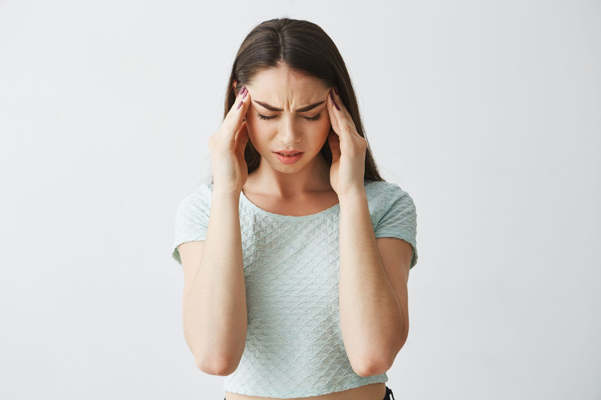Woman holding her temples with eyes closed, appearing stressed or in pain against a plain background