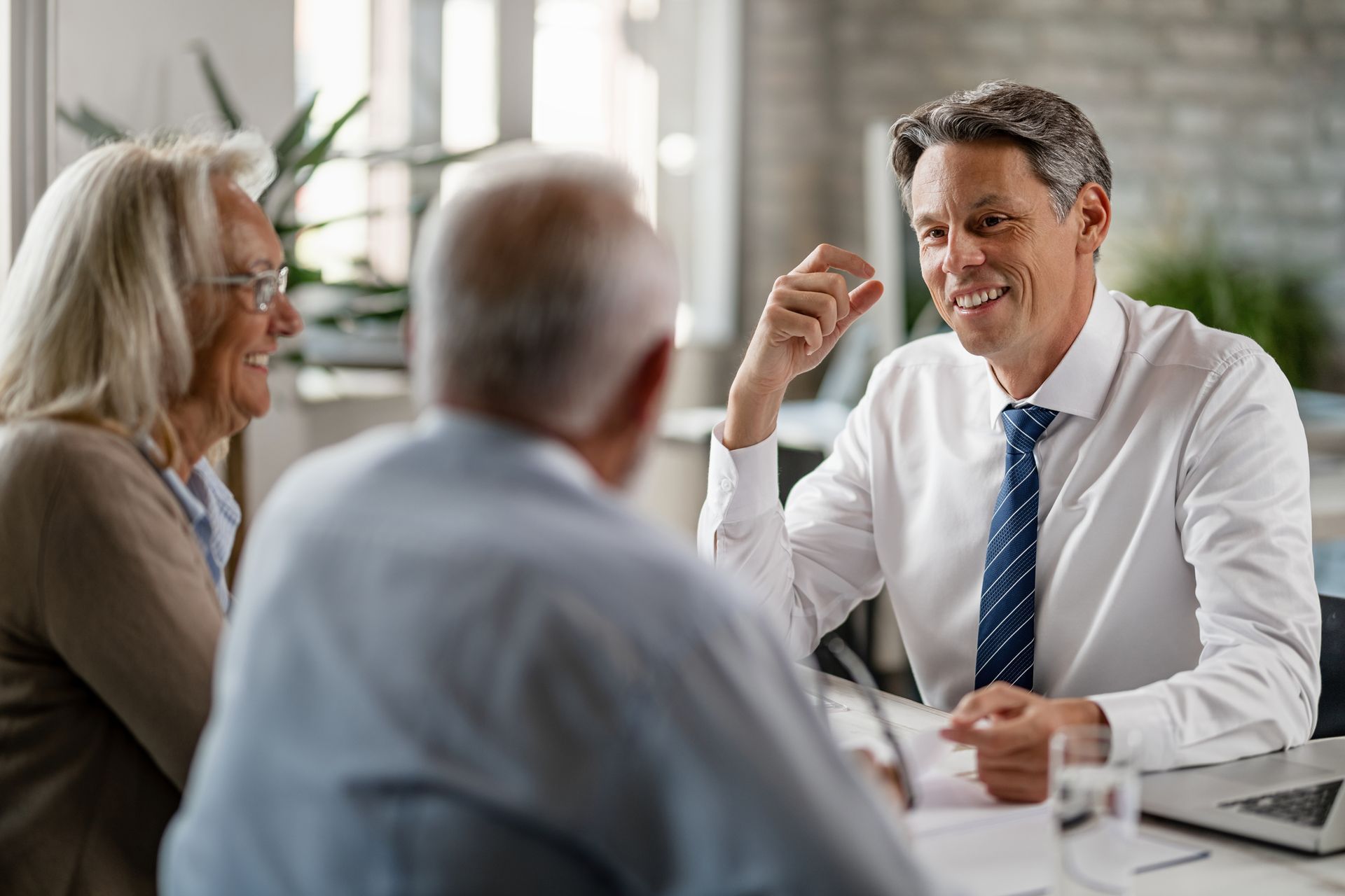 Financial advisor talking with a senior couple in an office; smiling, paperwork, laptop.
