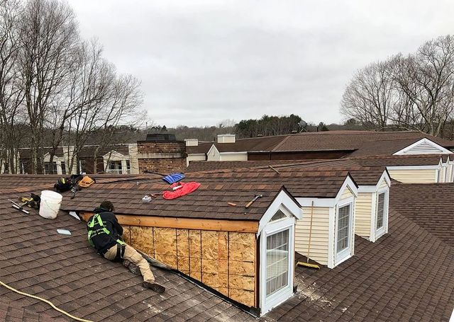 Roofers working on a brown shingle roof, near dormer windows, cloudy sky.