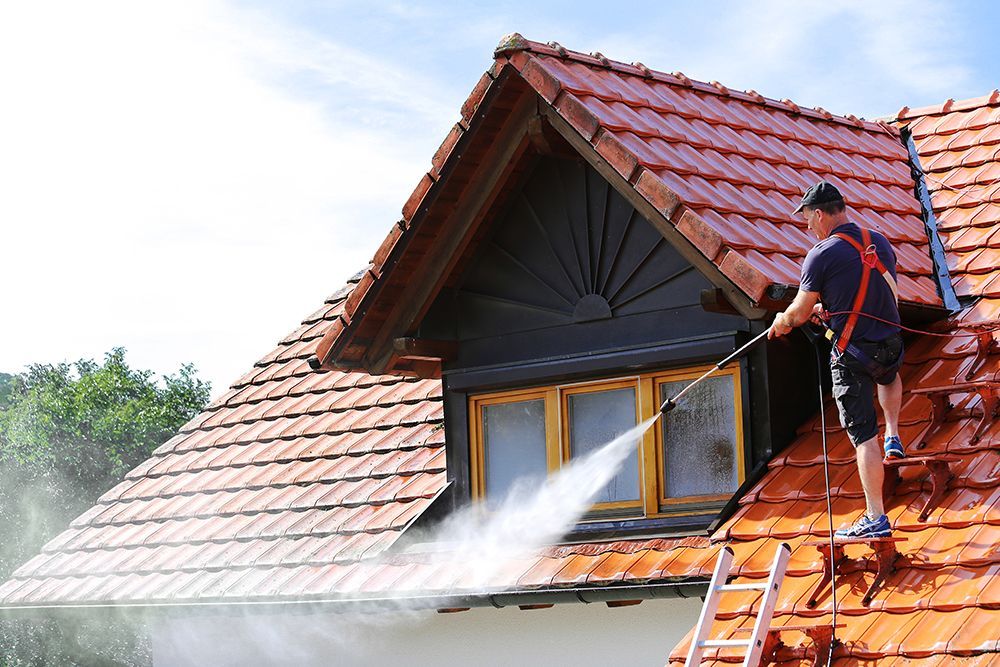Man pressure washing a red tiled roof, near a dormer window, on a sunny day.