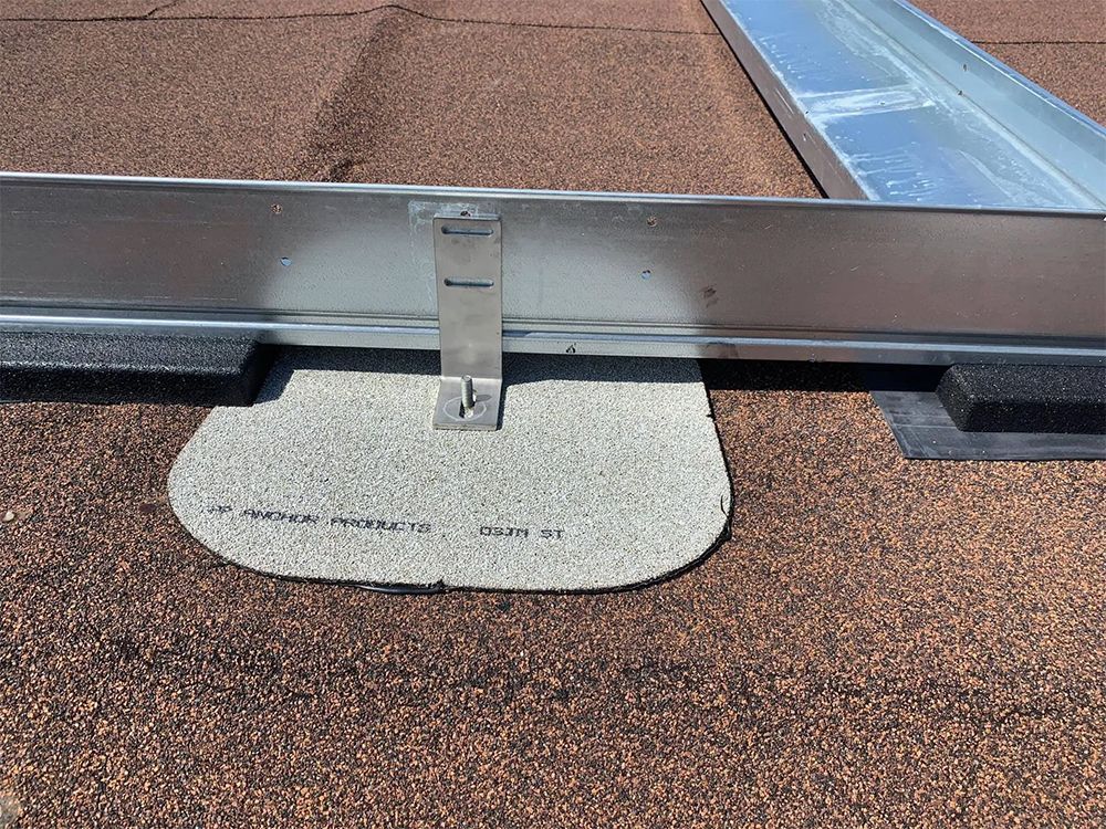 Close-up of a solar panel mount installed on a brown asphalt shingle roof.