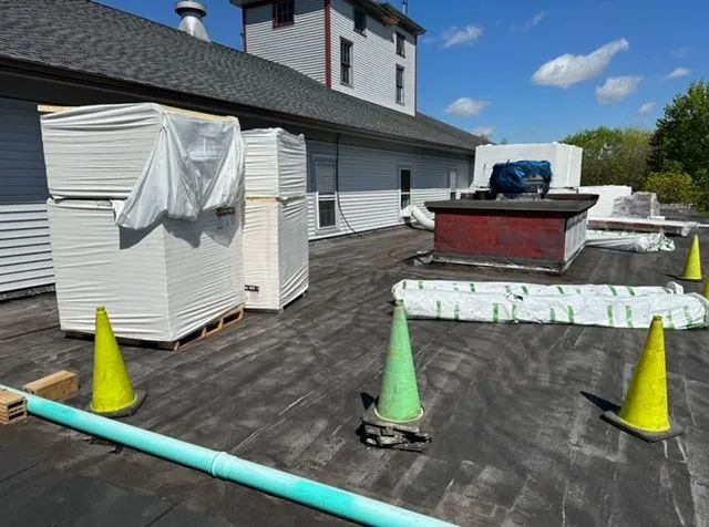Stacks of building materials on a flat roof, with safety cones and a blue pipe. The building is white.
