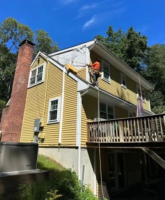Man on a roof repairing shingles on a yellow house with a brick chimney and wooden deck. Sunny day.
