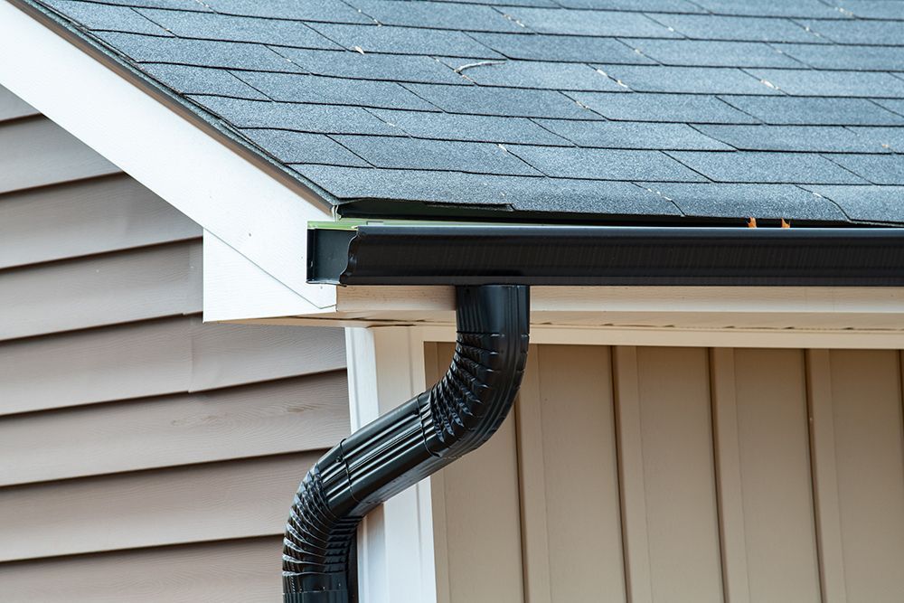 Black rain gutter attached to a roof with shingles and beige siding.