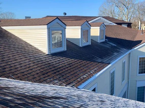 Brown shingled roof with three dormers, a light yellow exterior, and a snowy edge, set on a sunny day.