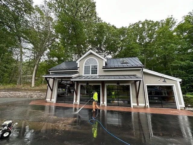 Person pressure washing a light-colored building with large glass garage doors, trees in the background.