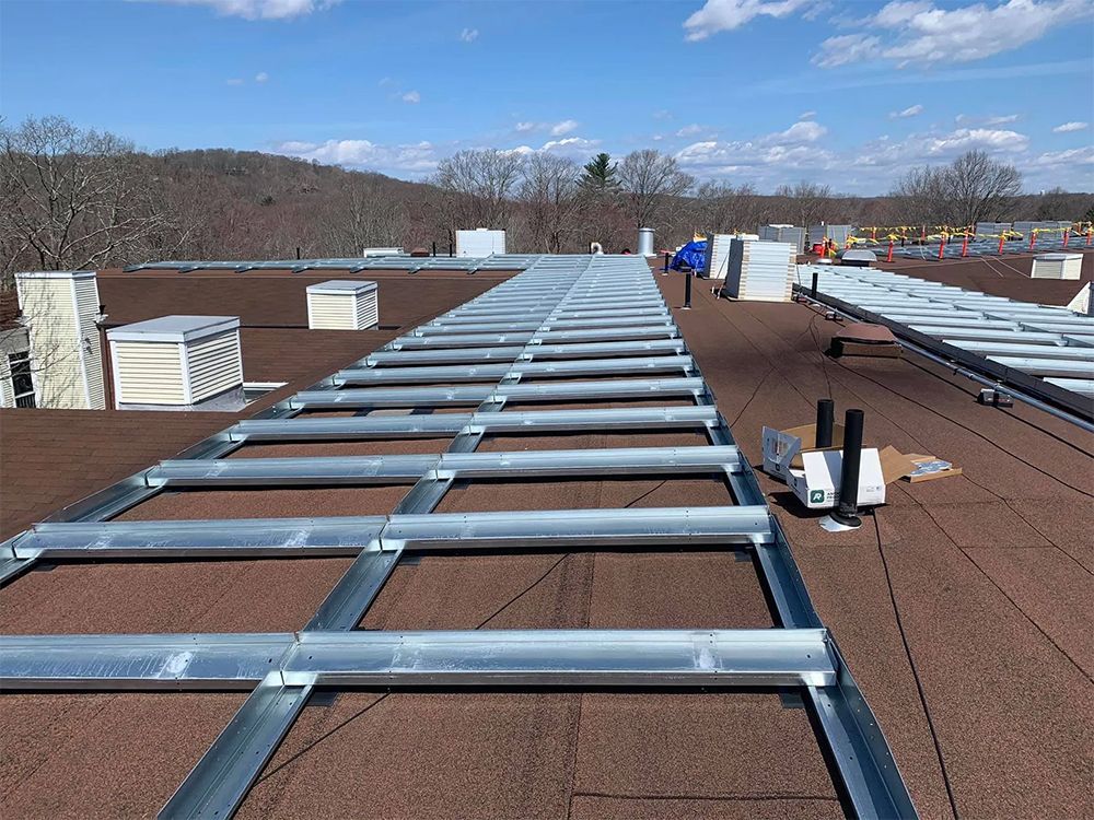 Solar panel racking installed on a flat, brown roof. A person walks between the rows. Blue sky, trees in the background.